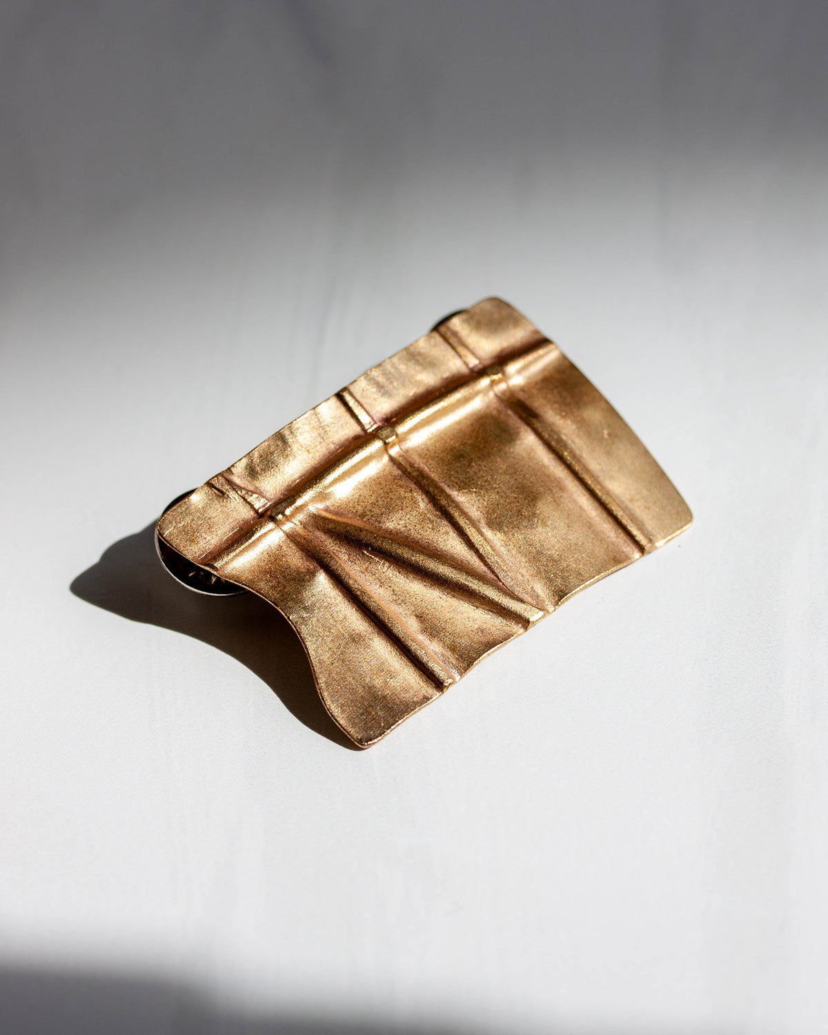 A brass brooch with a geometric design resembling folded metal segments, displayed on a two-tone background.