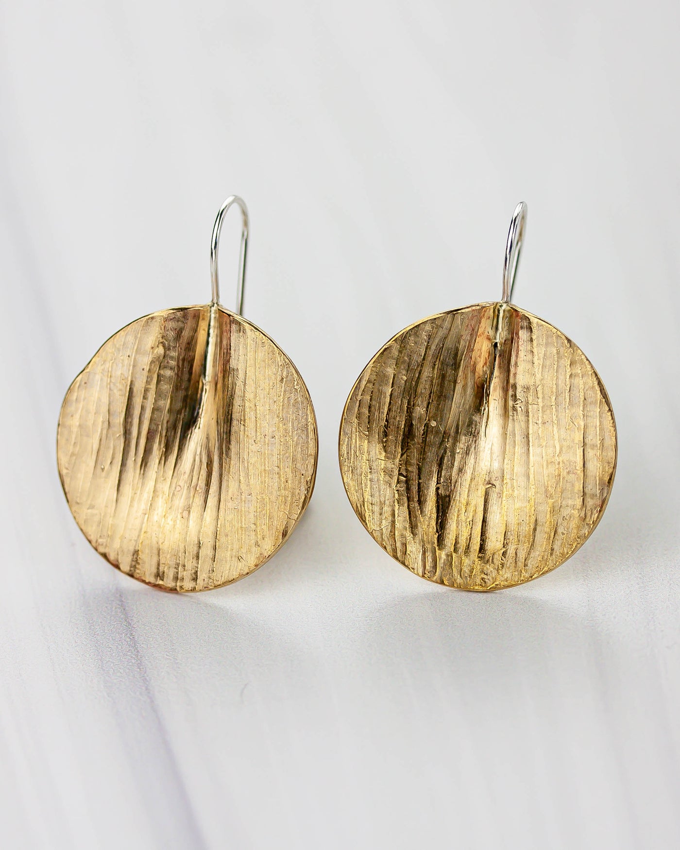 Golden bronze earrings on a white background. The shape is circular, lightly textured and folded to create a 3D effect.