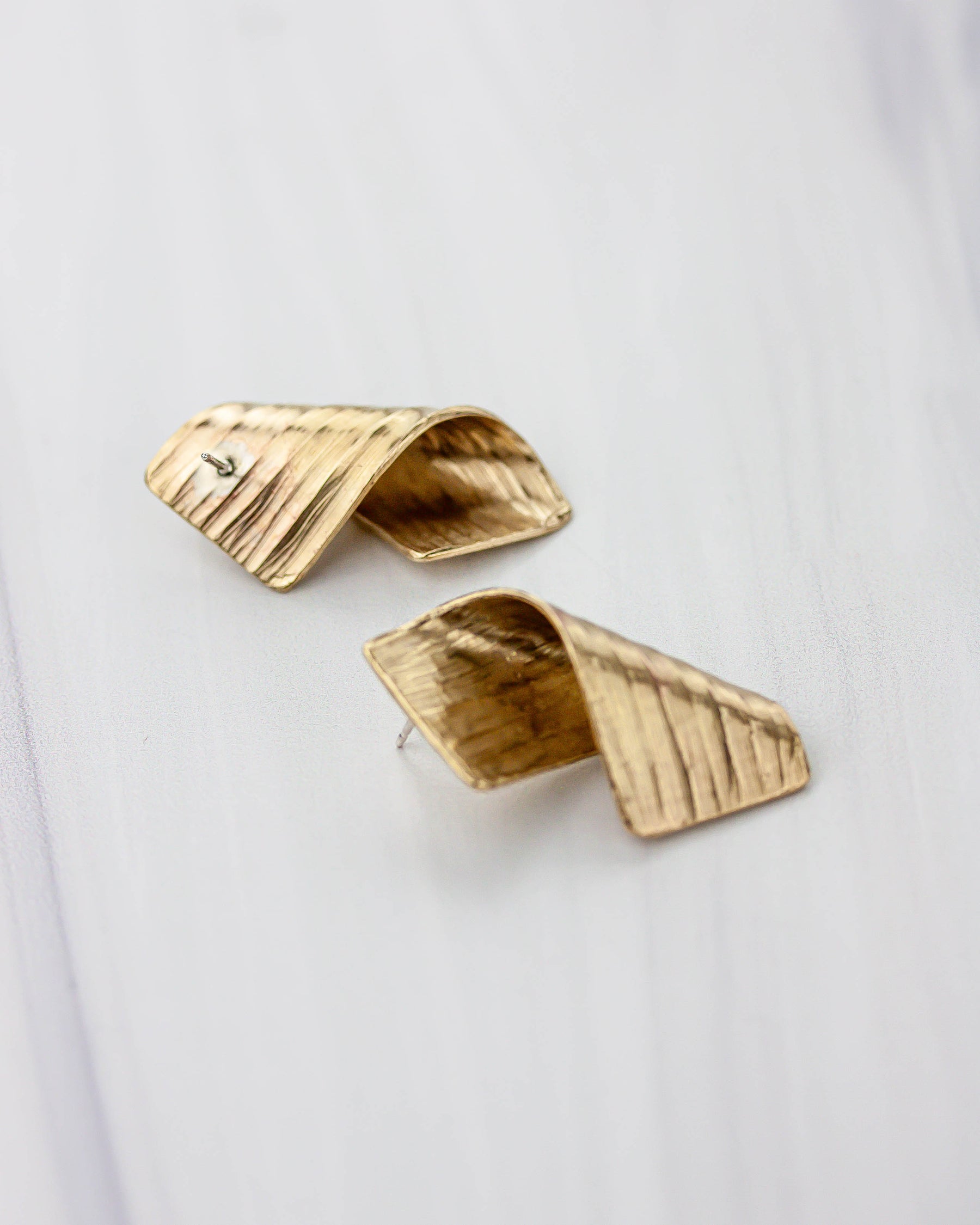 Golden bronze earrings on a white background. The shape is lightly textured and the metal is folded upon itself. They are flipped over so you can see the silver ear post.